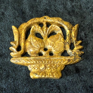 May include: A carved golden brown stone pendant depicting two birds in a basket. The birds are facing each other and have intricate details. The basket has a woven pattern.