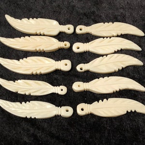 May include: Eight white carved bone feather charms with a hole at the top for hanging. The feathers are detailed with carved lines and have a smooth, polished finish.