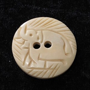 May include: A white, round button with a carved elephant design. The button has two holes for sewing.