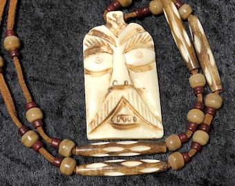 May include: A carved bone pendant with a face design hangs from a brown leather cord with brown and red beads. The pendant is attached to the cord with a metal loop.