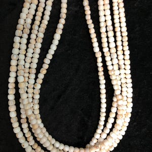 May include: Several strands of off-white, square-shaped beads are displayed against a black background. The beads have a slightly irregular shape and a subtle, natural variation in color, with hints of tan and cream. The strands are arranged in a cascading pattern.