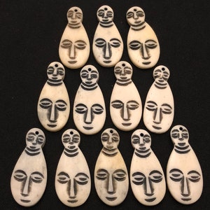 May include: Twelve white bone pendants with black face designs. Each pendant is shaped like a teardrop and has a different facial expression.