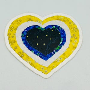 May include: A heart-shaped sticker with a layered design. The outer layer is yellow with a floral pattern, followed by a white layer, a blue layer with floral accents, and a black center with gold stars. The sticker has a white border.