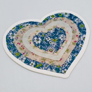 May include: A heart-shaped sticker with a white border. The design features layered hearts with floral patterns in shades of blue, pink, and white. The sticker has a slightly textured appearance.