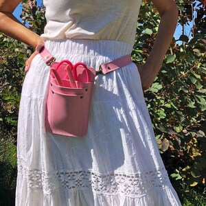 Pu&ograve; includere: Una cintura porta attrezzi in pelle rosa con una tasca per contenere le forbici. La cintura &egrave; indossata sopra una gonna bianca con pizzo.