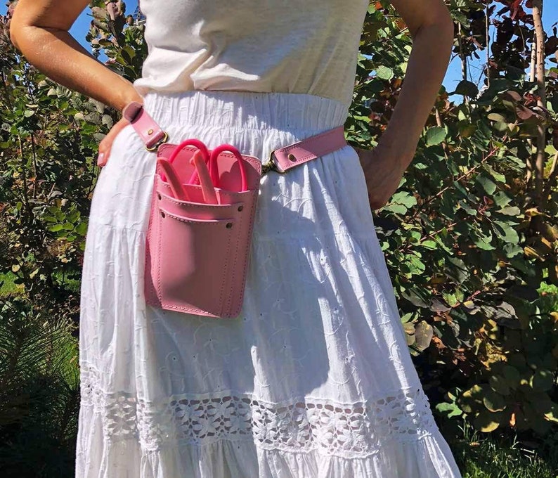 Pu&ograve; includere: Una cintura porta attrezzi in pelle rosa con una tasca per contenere le forbici. La cintura &egrave; indossata sopra una gonna bianca con pizzo.
