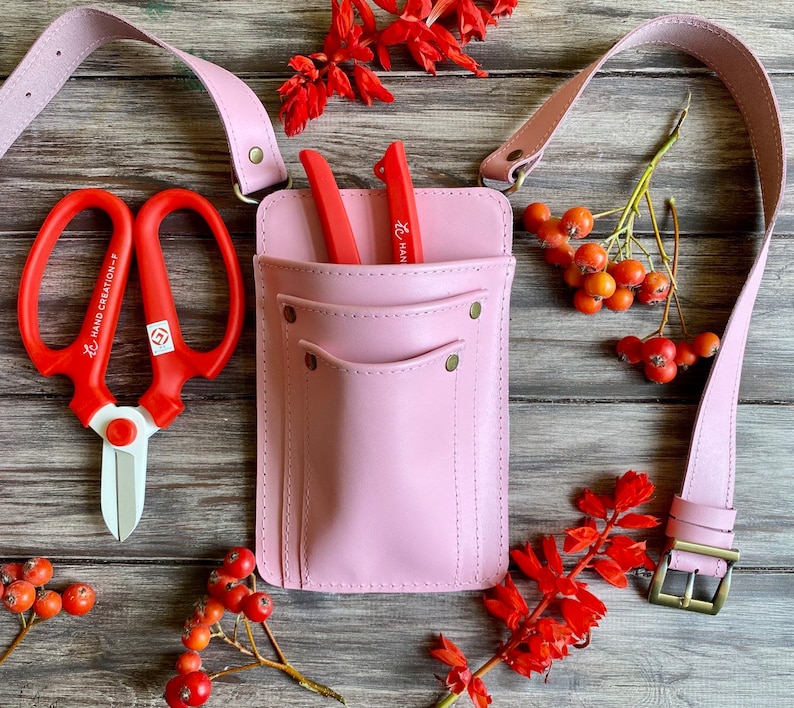 Pu&ograve; includere: Cintura da giardinaggio in pelle rosa con due tasche e una cinghia. La cintura ha due forbici da giardino rosse e un paio di forbici da potare rosse con manico bianco. Le forbici sono etichettate "Hand Creation" e "Han"