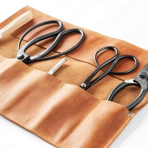 May include: A brown leather roll-up case with five slots for holding gardening tools. The case contains three pairs of black pruning shears and a small silver tool.