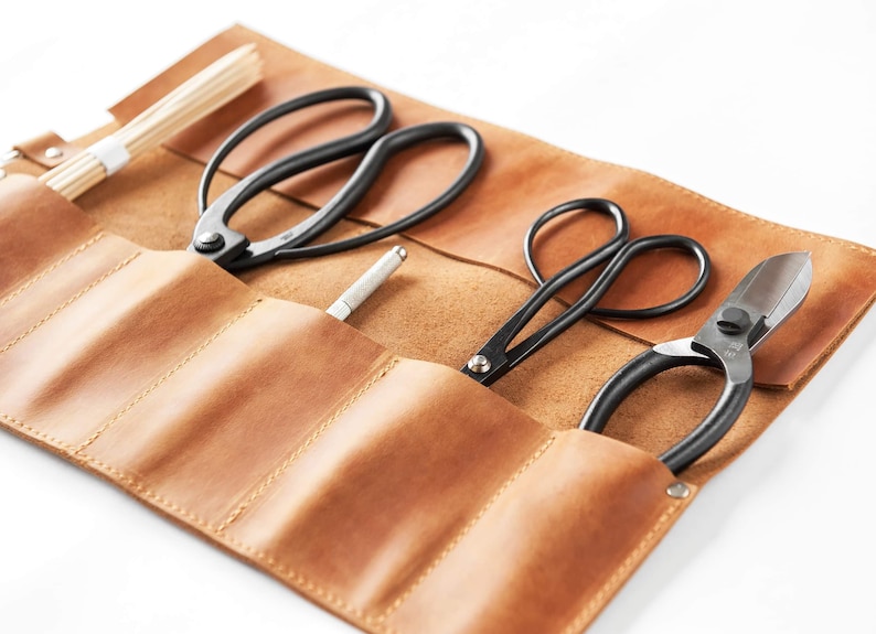 May include: A brown leather roll-up case with five slots for holding gardening tools. The case contains three pairs of black pruning shears and a small silver tool.
