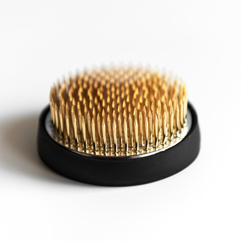 May include: A round, black floral frog with numerous gold-coloured pins. The pins are closely spaced and arranged in a circular pattern, creating a dense, textured surface. The frog sits on a white background.