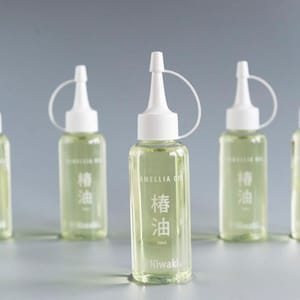 Könnte beinhalten: Fünf transparente Flaschen Kamelienöl mit weißen Verschlüssen und Düsen. Die Flaschen sind mit einer hellgrünen Flüssigkeit gefüllt und tragen japanische Schriftzeichen und die Worte "Camellia Oil". Die Flaschen sind 120 ml.