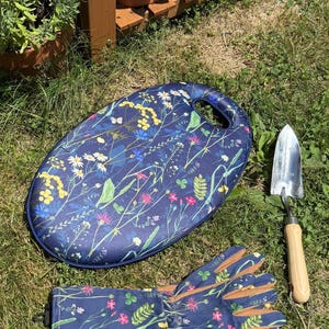 May include: A navy blue garden kneeling pad with a colorful wildflower print, a pair of matching long-cuffed gardening gloves with tan palms, and a metal hand trowel with a wooden handle, all featuring a floral pattern of daisies and cornflowers.