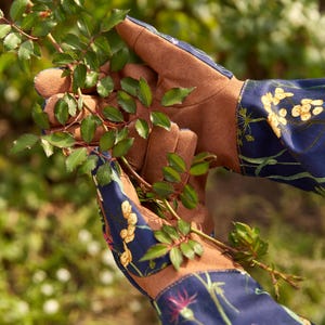 May include: A pair of gardening gloves with brown leather palms and blue cotton backs. The cotton fabric has a floral print with yellow, green, and pink flowers. The gloves are holding a green plant stem.