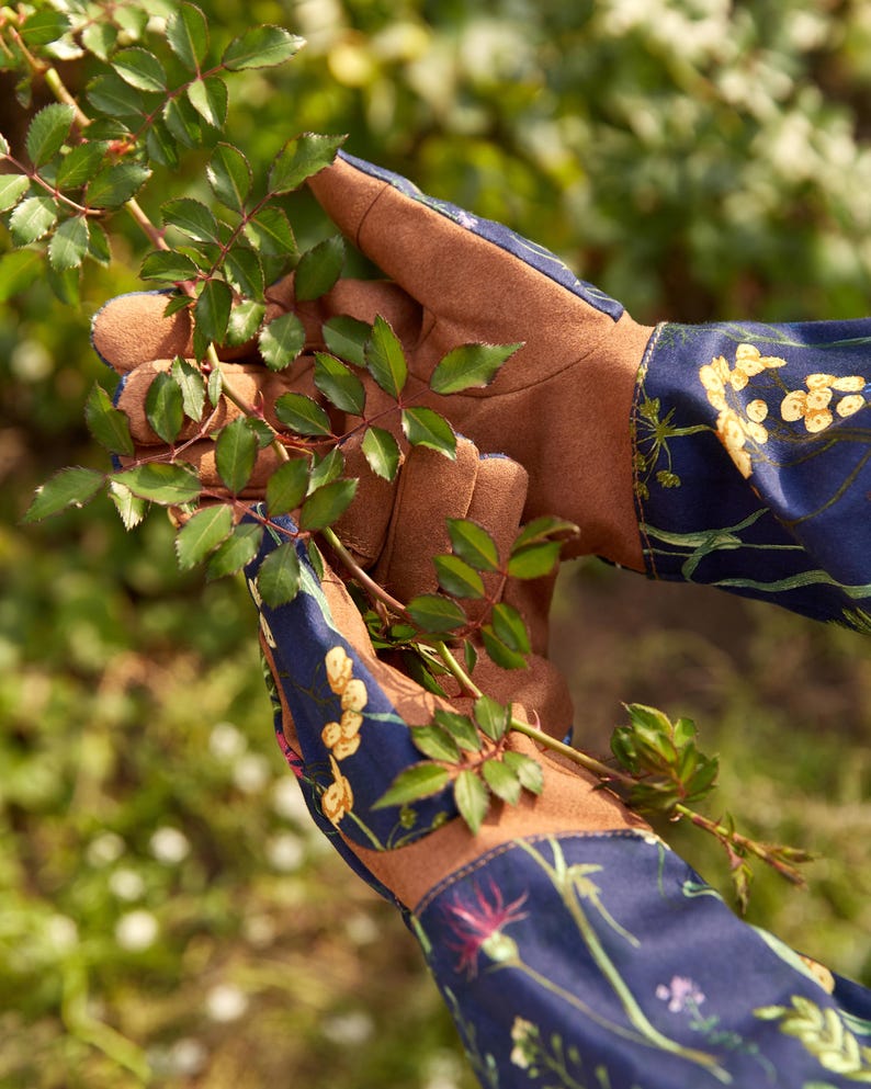 May include: A pair of gardening gloves with brown leather palms and blue cotton backs. The cotton fabric has a floral print with yellow, green, and pink flowers. The gloves are holding a green plant stem.