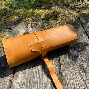 May include: A rolled-up brown leather case with a strap and buckle closure. The case is lying on a wooden surface.