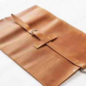 May include: A brown leather roll-up case with multiple slots for holding tools or other items. The case has a strap with a metal buckle and a metal ring.