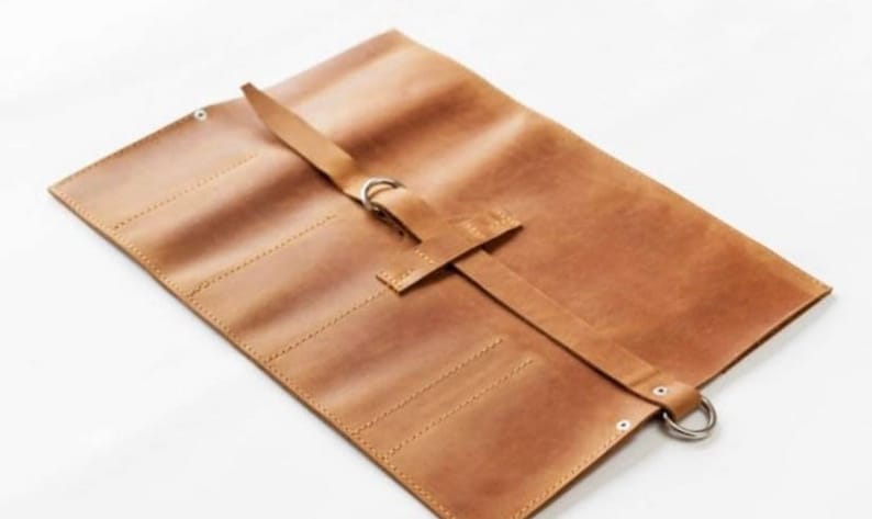 May include: A brown leather roll-up case with multiple slots for holding tools or other items. The case has a strap with a metal buckle and a metal ring.