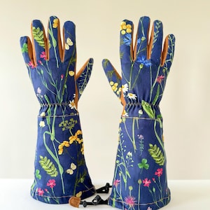 May include: A pair of blue gardening gloves with a floral print featuring yellow, pink, and white flowers, green leaves, and butterflies. The gloves have brown leather palms and a black adjustable strap.