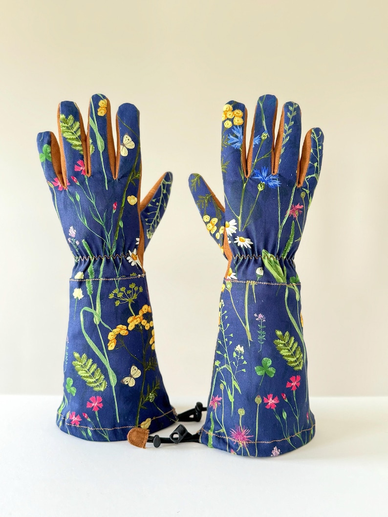 May include: A pair of blue gardening gloves with a floral print featuring yellow, pink, and white flowers, green leaves, and butterflies. The gloves have brown leather palms and a black adjustable strap.