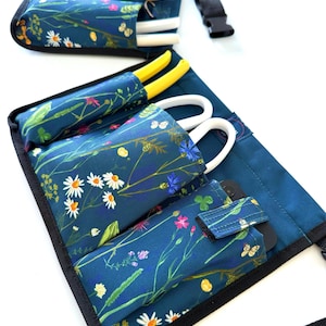 May include: A teal gardening tool belt with multiple pockets and a black adjustable strap. The belt is decorated with a floral print of white daisies, pink flowers, and green leaves.