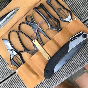 May include: A brown leather roll-up case with a variety of gardening tools, including pruning shears, a saw, and a knife. The tools are arranged in a row and secured with a strap.