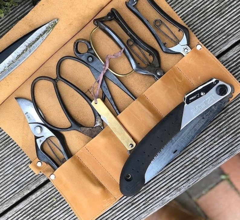 May include: A brown leather roll-up case with a variety of gardening tools, including pruning shears, a saw, and a knife. The tools are arranged in a row and secured with a strap.
