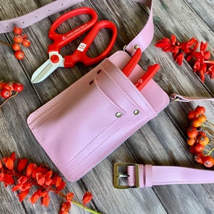 Pu&ograve; includere: Una cintura porta attrezzi in pelle rosa con una tasca e una fibbia. La cintura ha un paio di forbici da potatura rosse e bianche e un paio di forbici da potatura rosse.