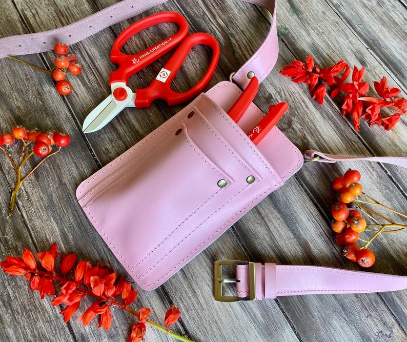 Pu&ograve; includere: Una cintura porta attrezzi in pelle rosa con una tasca e una fibbia. La cintura ha un paio di forbici da potatura rosse e bianche e un paio di forbici da potatura rosse.
