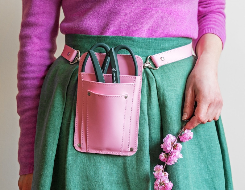 Pu&ograve; includere: Una cintura da giardinaggio in pelle rosa con una tasca e un paio di forbici da giardino nere. La cintura &egrave; indossata sopra una gonna verde.