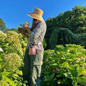 Pu&ograve; includere: Una donna con un cappello di paglia e una camicia a stampa floreale &egrave; in piedi in un giardino. Tiene una borsa porta attrezzi da giardinaggio rosa con un paio di forbici all'interno. La borsa &egrave; attaccata a una cintura intorno alla sua vita. Il giardino &egrave; pieno di piante verdi e fiori rosa.