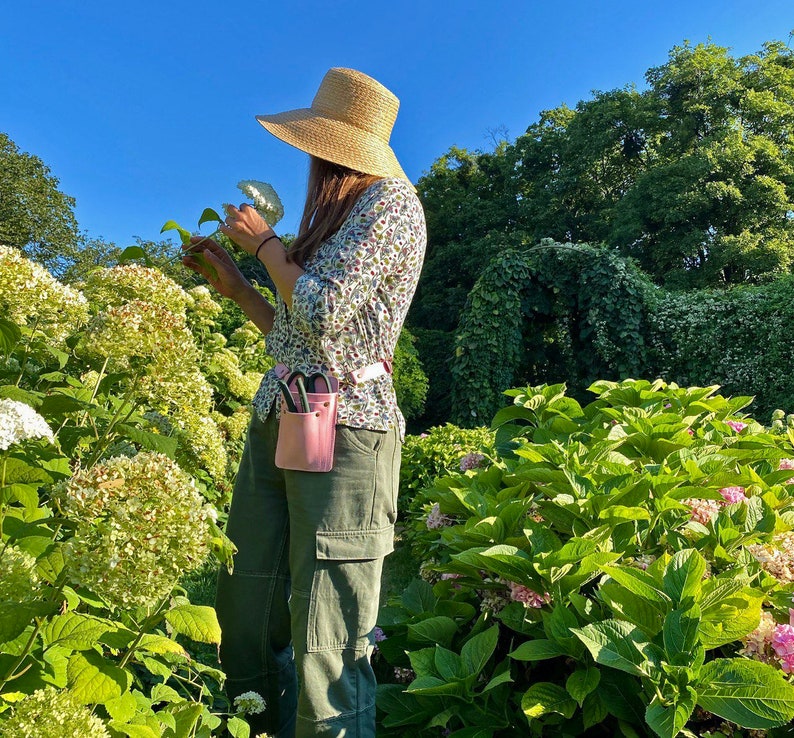 Pu&ograve; includere: Una donna con un cappello di paglia e una camicia a stampa floreale &egrave; in piedi in un giardino. Tiene una borsa porta attrezzi da giardinaggio rosa con un paio di forbici all'interno. La borsa &egrave; attaccata a una cintura intorno alla sua vita. Il giardino &egrave; pieno di piante verdi e fiori rosa.