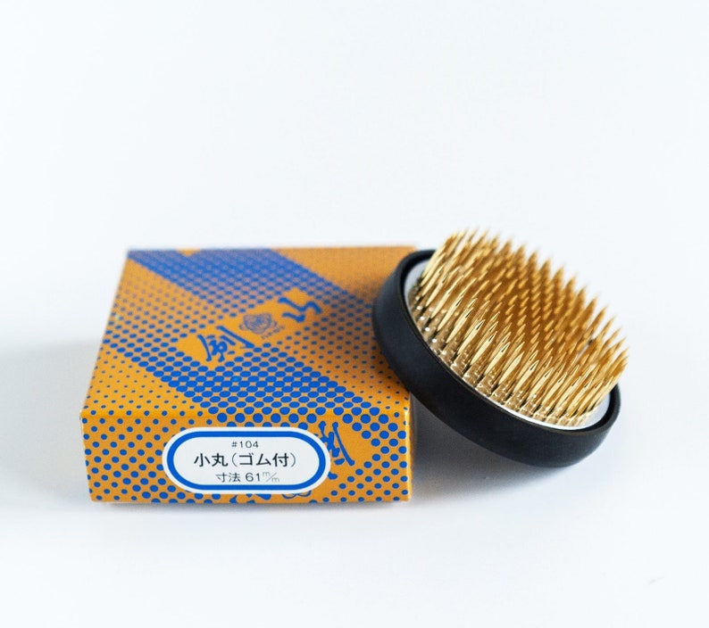 May include: A black and gold flower frog, or kenzan, with a box. The flower frog has a black base and gold pins. The box is orange with a blue dot pattern and Japanese text.