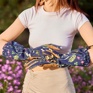 May include: A pair of blue gardening gloves with a floral print. The gloves have a brown leather palm and are being worn by a person with a white shirt and a beige skirt.