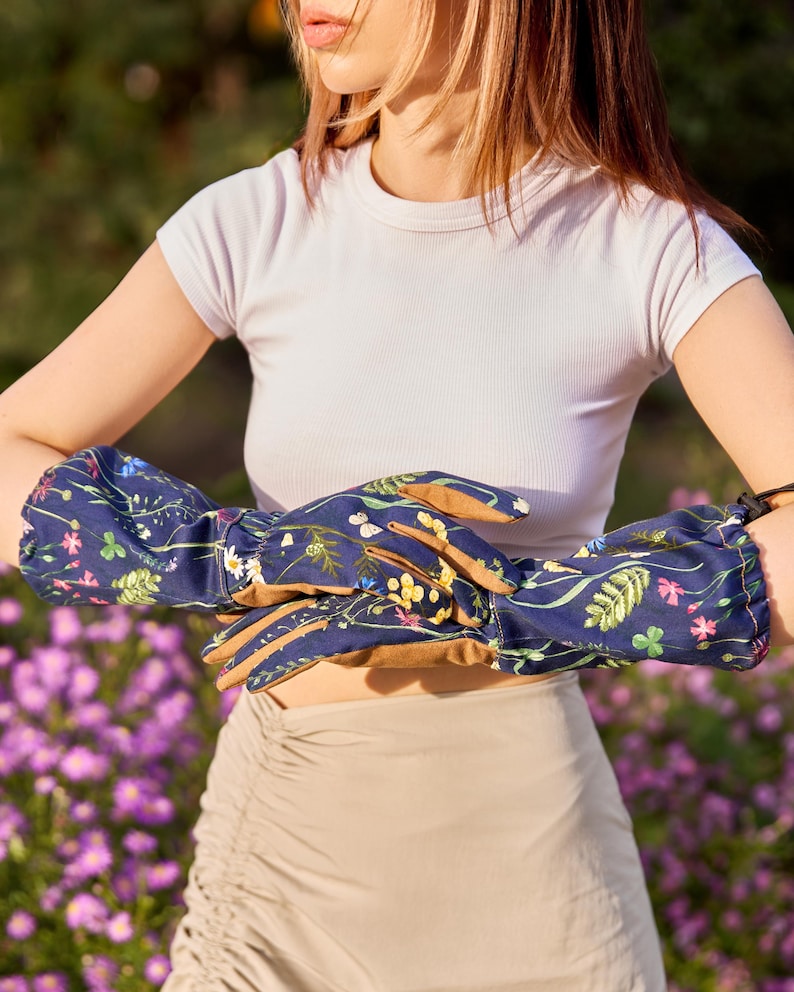 May include: A pair of blue gardening gloves with a floral print. The gloves have a brown leather palm and are being worn by a person with a white shirt and a beige skirt.