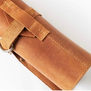 May include: A brown leather roll with a strap and buckle closure. The roll has a silver metal ring on the side.