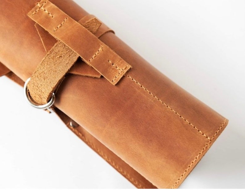 May include: A brown leather roll with a strap and buckle closure. The roll has a silver metal ring on the side.
