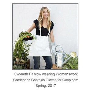 May include: Gwyneth Paltrow wearing a white apron and black top, holding a bowl of vegetables and a watering can. The text "Gwyneth Paltrow wearing Womanwork Gardener's Goatskin Gloves for Goop.com Spring, 2017" is below the image.