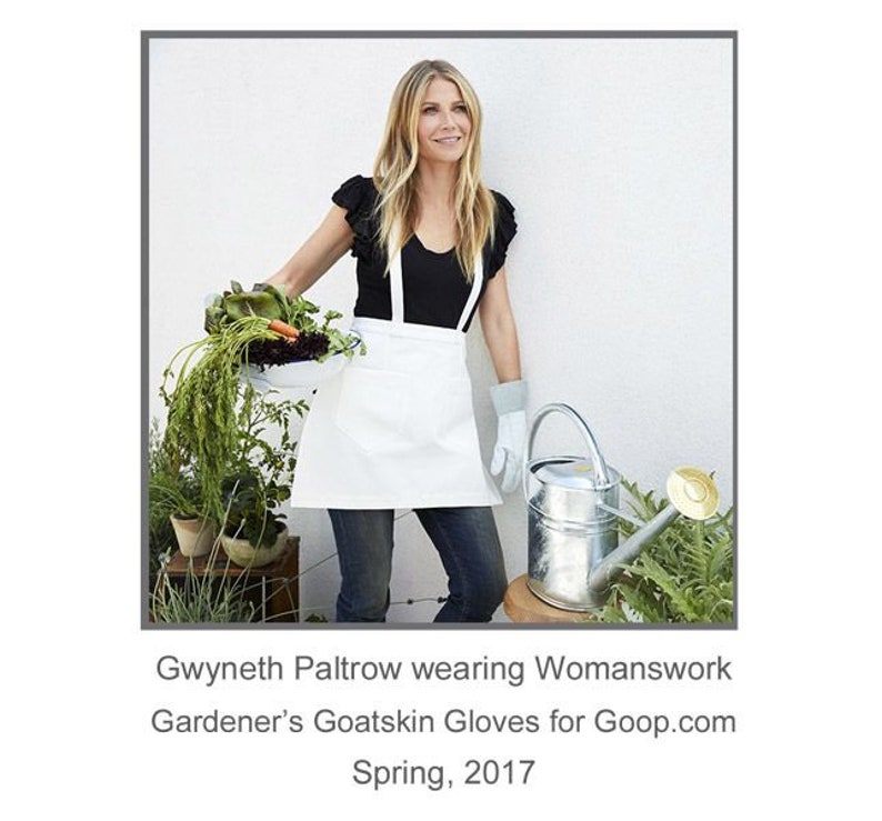 May include: Gwyneth Paltrow wearing a white apron and black top, holding a bowl of vegetables and a watering can. The text "Gwyneth Paltrow wearing Womanwork Gardener's Goatskin Gloves for Goop.com Spring, 2017" is below the image.
