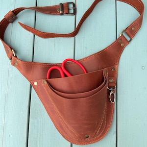 May include: A brown leather tool belt with multiple pockets and a belt strap. Red scissors are visible in one of the pockets. The belt has brass-colored rivets and a metal clasp.
