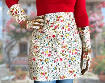 May include: A white apron with a floral print featuring small pink, red, and yellow flowers. The apron has a brown leather trim and is worn over a red shirt and blue jeans.