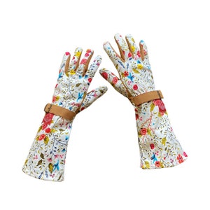Op de afbeelding: Een paar witte tuinhandschoenen met een bloemenprint en bruine leren banden. De handschoenen hebben een lange manchet die voorbij de pols reikt.