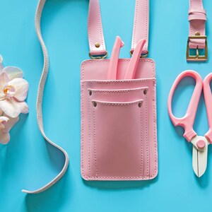 Pu&ograve; includere: Una cintura portautensili in pelle rosa con una tasca e due strumenti all'interno. La cintura ha una lunga cinghia e una fibbia. Ci sono delle forbici rosa e un paio di cesoie rosa su uno sfondo blu.