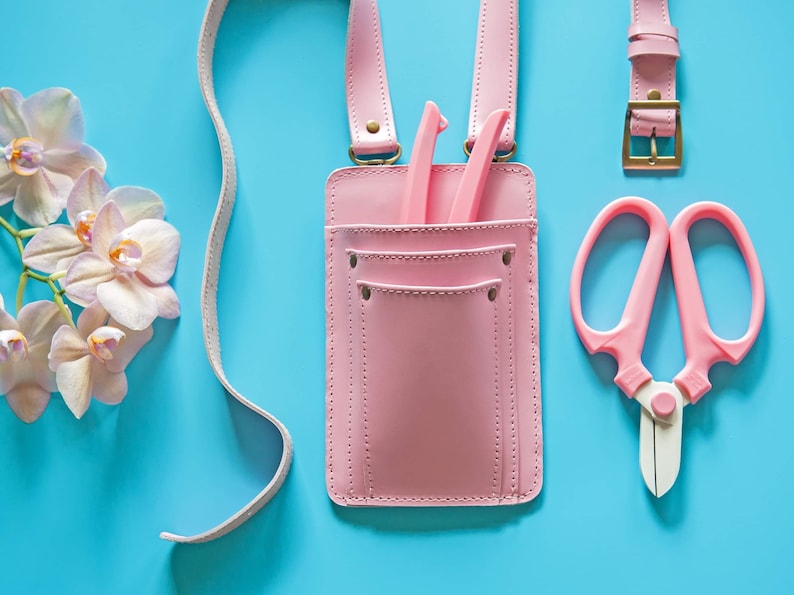 Pu&ograve; includere: Una cintura portautensili in pelle rosa con una tasca e due strumenti all'interno. La cintura ha una lunga cinghia e una fibbia. Ci sono delle forbici rosa e un paio di cesoie rosa su uno sfondo blu.