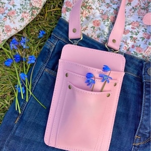 Pu&ograve; includere: Una borsa a tracolla in pelle rosa con una sola tasca e una chiusura a lembo. La borsa &egrave; indossata sopra un paio di jeans blu e una camicia floreale. Dei fiori blu sono infilati nella tasca.