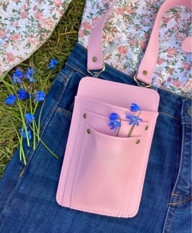 Pu&ograve; includere: Una borsa a tracolla in pelle rosa con una sola tasca e una chiusura a lembo. La borsa &egrave; indossata sopra un paio di jeans blu e una camicia floreale. Dei fiori blu sono infilati nella tasca.