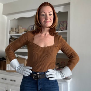 May include: A woman wearing a brown ribbed sweater with a square neckline, white leather gloves, a black belt, and blue jeans. She is standing in front of a white cabinet with shelves.