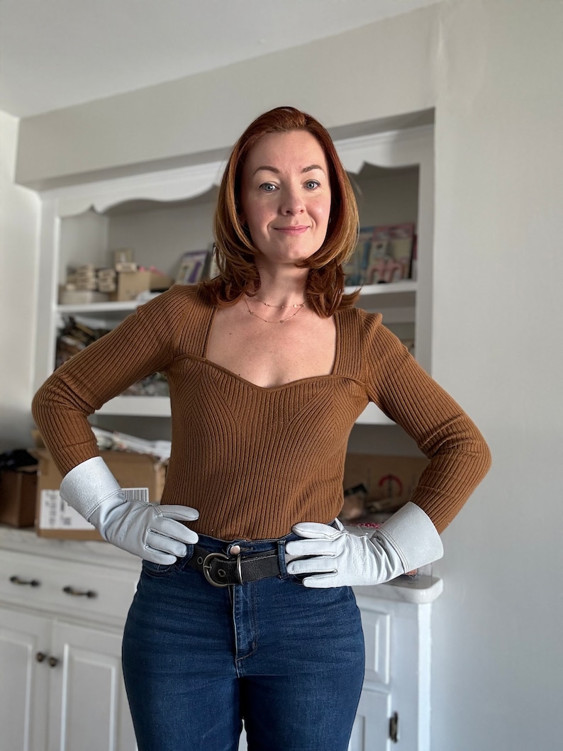 May include: A woman wearing a brown ribbed sweater with a square neckline, white leather gloves, a black belt, and blue jeans. She is standing in front of a white cabinet with shelves.