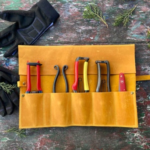 May include: A tan leather roll-up case with six slots for holding gardening tools. The case is open and shows five tools, including a pair of red pruning shears, a pair of yellow pruning shears, and three pairs of black pruning shears. A small red pocket knife is also visible in the case.