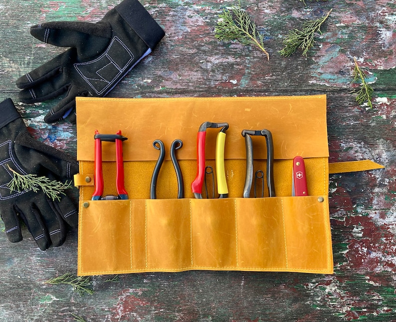 May include: A tan leather roll-up case with six slots for holding gardening tools. The case is open and shows five tools, including a pair of red pruning shears, a pair of yellow pruning shears, and three pairs of black pruning shears. A small red pocket knife is also visible in the case.