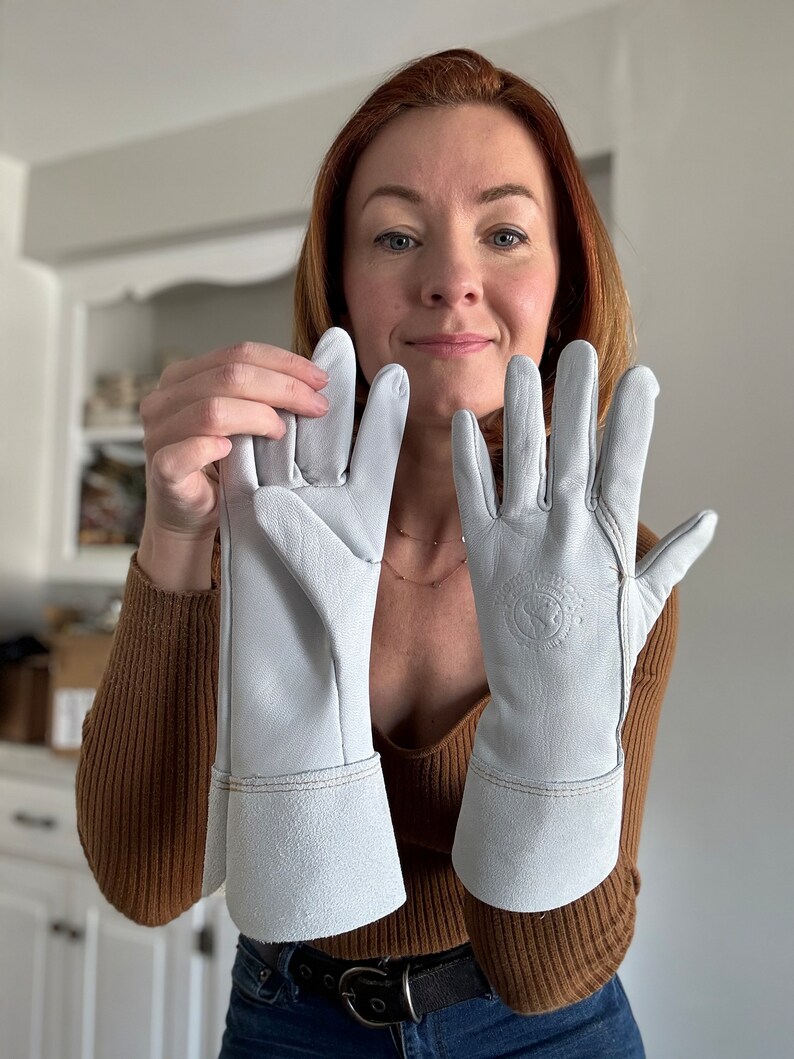 May include: A pair of white leather gloves with a cuff. The gloves have a logo on the back of the right glove.
